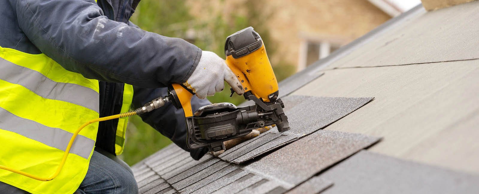 Unrecognizable roofer worker in special protective work wear and gloves, using air or pneumatic nail gun and installing asphalt or bitumen shingle on top of the new roof under construction residential building